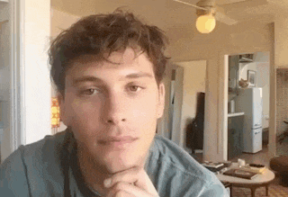a man in a blue shirt is sitting in a living room with his hand on his chin .