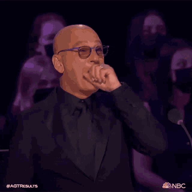 a bald man wearing glasses and a black suit is sitting in front of a microphone with his hand to his mouth .