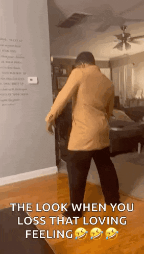 a man in a brown jacket is standing in a living room with a ceiling fan .