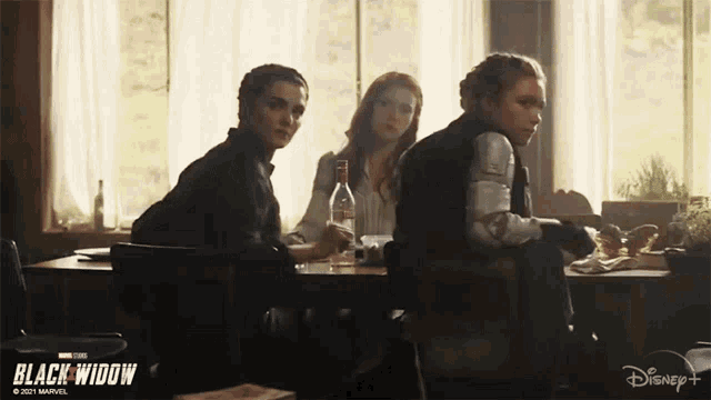 three women are sitting at a table in front of a window with a poster for black widow on the wall