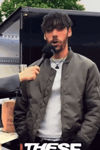 a man in a bomber jacket is being interviewed with a microphone and the word these is on the bottom right