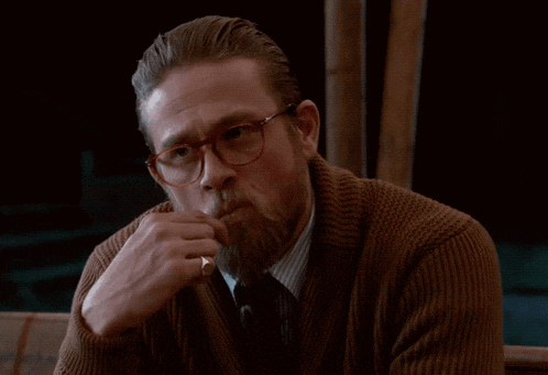 a man with a beard wearing glasses and a brown sweater is smoking a cigarette