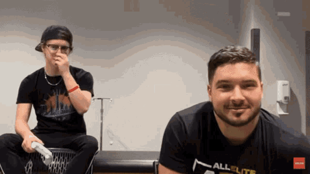 a man wearing a black shirt that says all elite is sitting next to another man