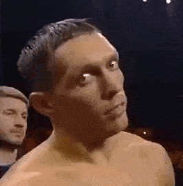 a man without a shirt is standing in a dark room with a man in the background .
