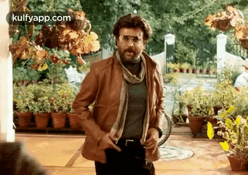 a man in a brown leather jacket and scarf is standing in front of a row of potted plants .