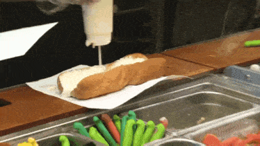 a hot dog with mayonnaise being poured on it sits on a counter