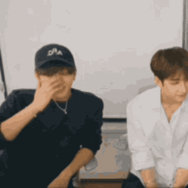 two men are sitting next to each other in a room . one of the men is wearing a baseball cap .