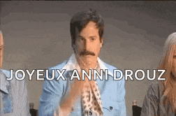 a man with a mustache is sitting in front of a group of people with the words joyeux anni drouz written on the screen .