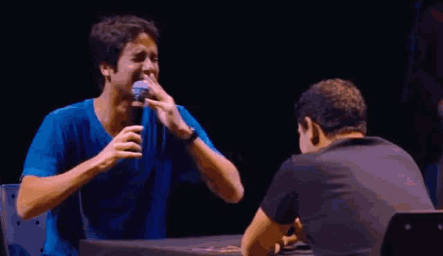 a man in a blue shirt is crying while talking into a microphone while another man looks on .