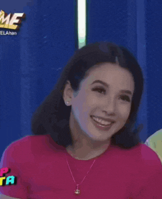 a woman wearing a pink shirt and a necklace smiles in front of a sign that says me