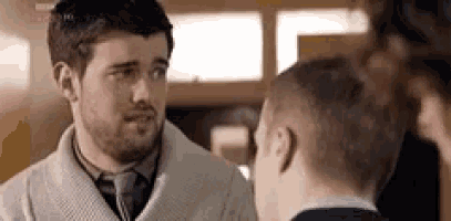 a man with a beard and tie is talking to another man in a room .