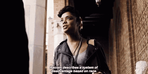 a woman in a black leather jacket is standing in front of a building and talking about racism .