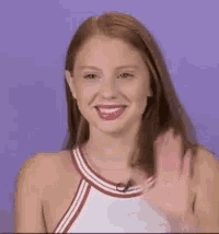 a woman is smiling and waving at the camera while wearing a white top .