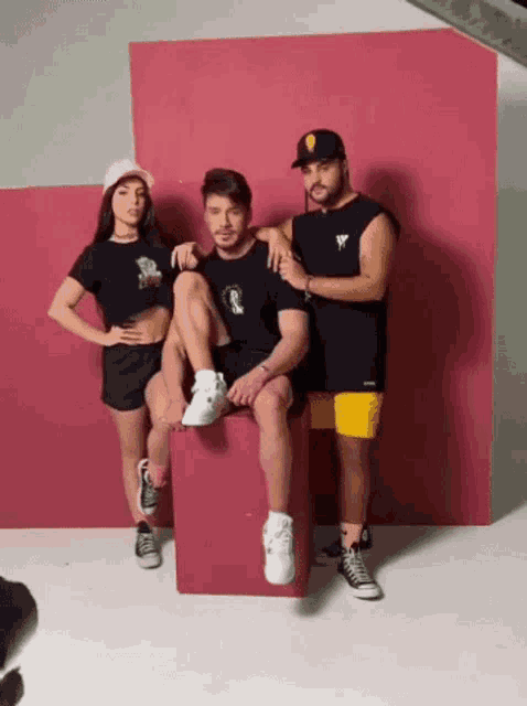 two men and a woman are posing for a picture in front of a pink wall . the woman is wearing a pink hat .