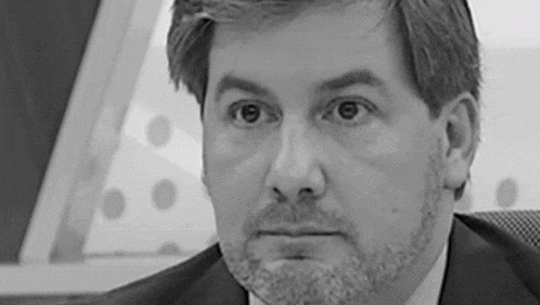 a man with a beard and a suit is sitting in a chair and looking at the camera in a black and white photo .