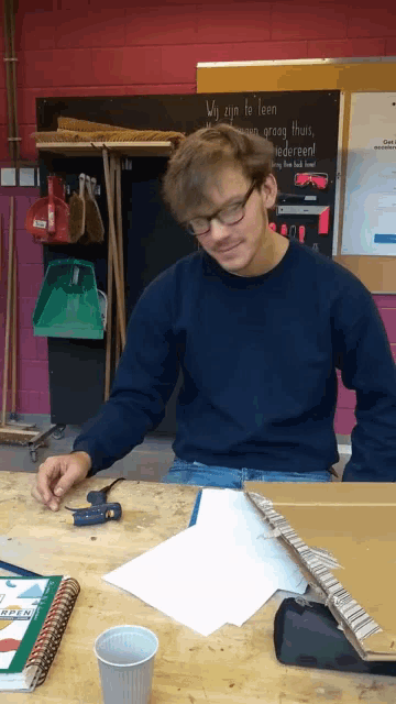 a man in a blue sweater sits at a table in front of a chalkboard that says wij zien to leen