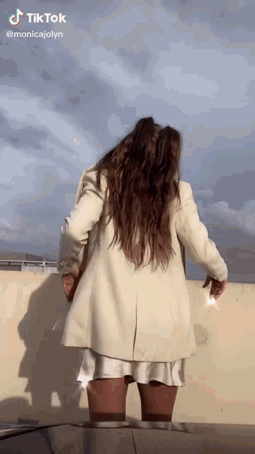 a woman in a white coat and white skirt is standing on a roof with a cloudy sky behind her