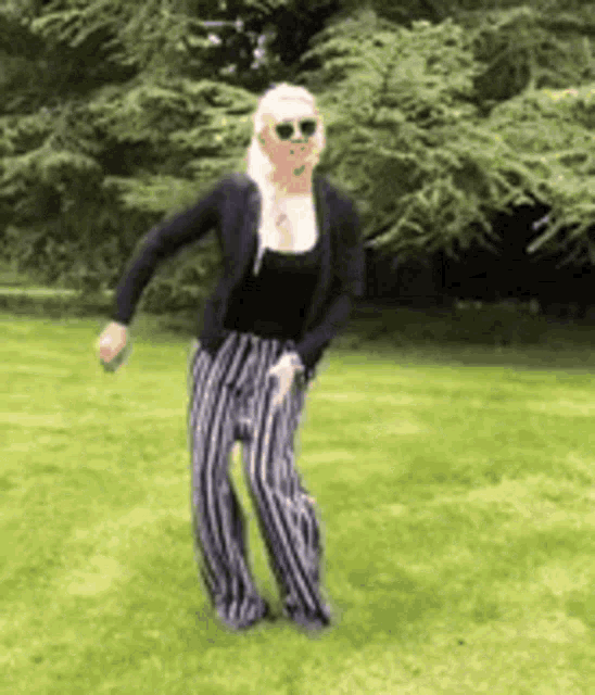 a woman wearing sunglasses and striped pants is dancing in a grassy field .