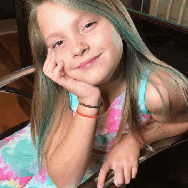 a young girl with green hair is sitting in a chair with her hand on her chin