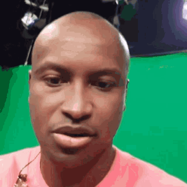 a bald man wearing a pink shirt is looking at the camera in front of a green screen .