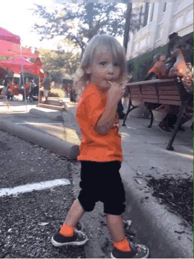 a little boy in an orange shirt and black shorts is walking down the street