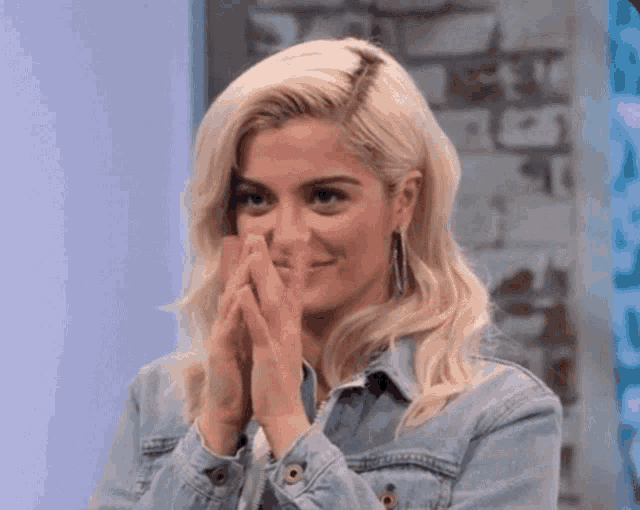 a woman with blonde hair is wearing a denim jacket and covering her face with her hands