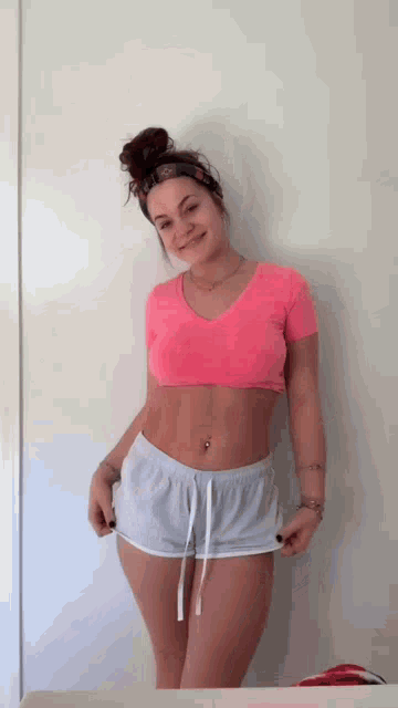 a woman in a pink crop top and white shorts is standing in front of a wall .