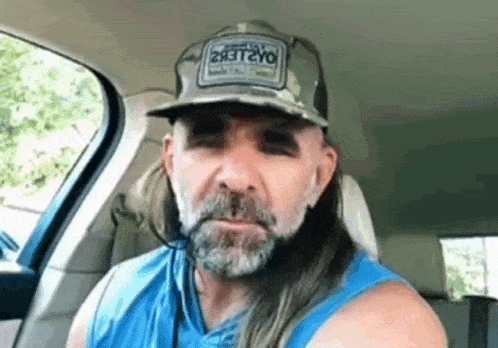 a man with long hair and a beard is sitting in a car wearing a hat .
