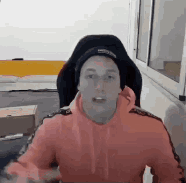 a man in a pink hoodie and black hat is sitting in a chair in front of a window .