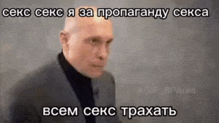 a bald man in a suit and black turtleneck is standing in front of a wall .