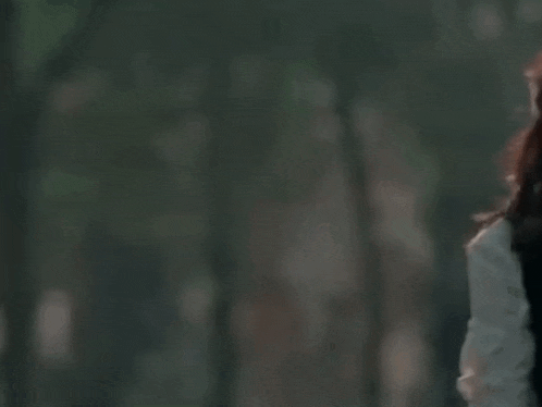 a woman with long red hair is standing with her arms outstretched in front of a forest .