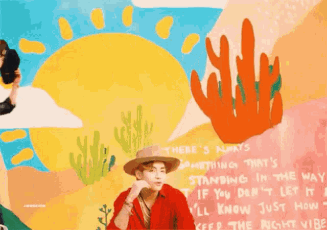 a man in a cowboy hat is standing in front of a wall with a painting of a sun and cactus .