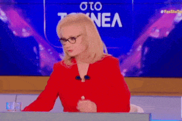 a woman wearing glasses and a red jacket is sitting at a desk in front of a screen that says to nea on it