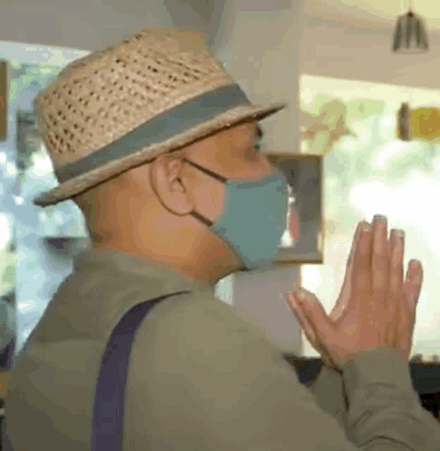 a man wearing a straw hat and a face mask is praying with his hands together .