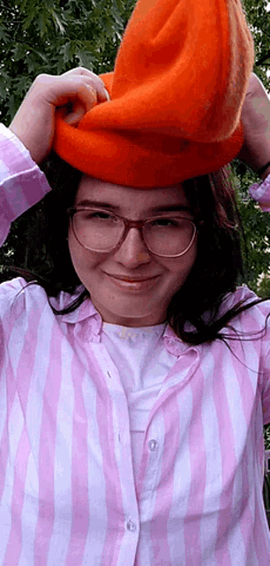 a girl wearing glasses and a pink and white striped shirt is holding an orange hat on her head