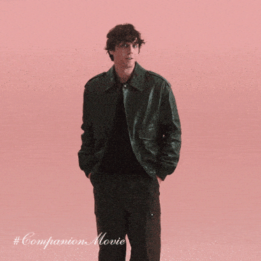 a man in a green leather jacket stands in front of a pink background with # companion movie written below him