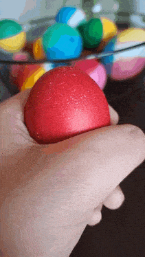 a hand is holding a red ball in front of a bowl of balls