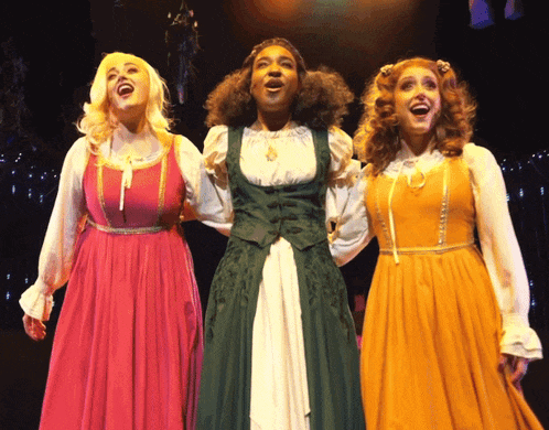 three women are singing on a stage and one is wearing a green vest