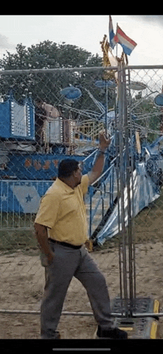a man in a yellow shirt is standing next to a chain link fence in front of a carnival ride .