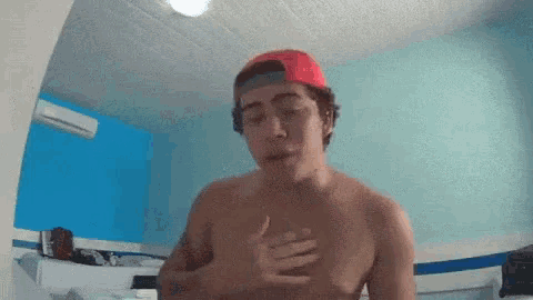 a shirtless man wearing a red hat is holding his chest in a room .
