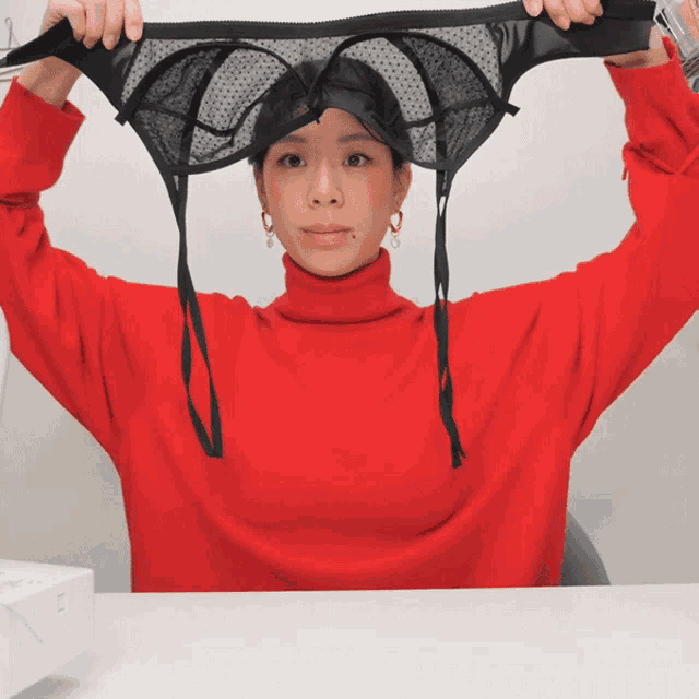 a woman in a red turtleneck is holding up a black bra