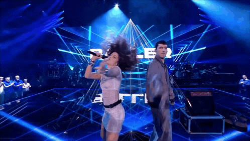 a woman sings into a microphone while a man stands behind her on a stage with a pyramid in the background