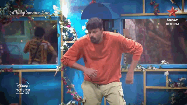 a man in an orange shirt is dancing in front of a blue wall with flowers and the words disney + hotstar on the bottom