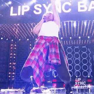 a man in a plaid shirt is singing into a microphone on a stage in front of a sign that says lip sync