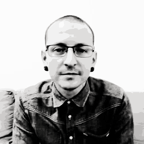 a man wearing glasses and a denim shirt is sitting on a couch in a black and white photo
