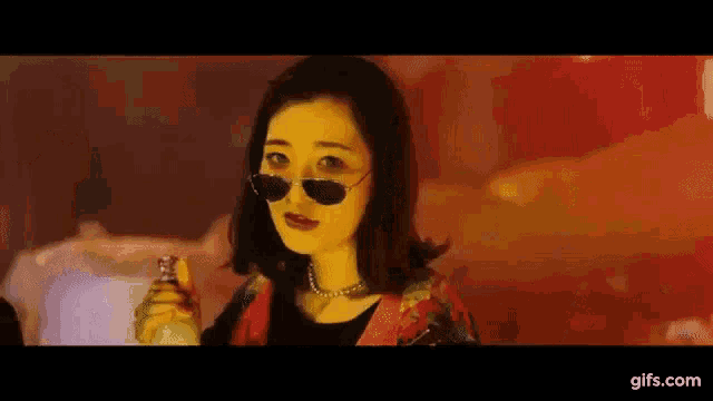 a woman wearing sunglasses and a necklace is holding a bottle of perfume .