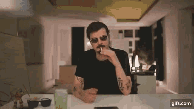 a man with a mustache and sunglasses is sitting at a table smoking a cigarette