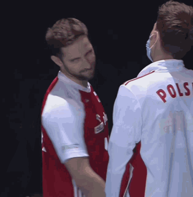 a man wearing a red orlen plus 12 jersey talks to another man