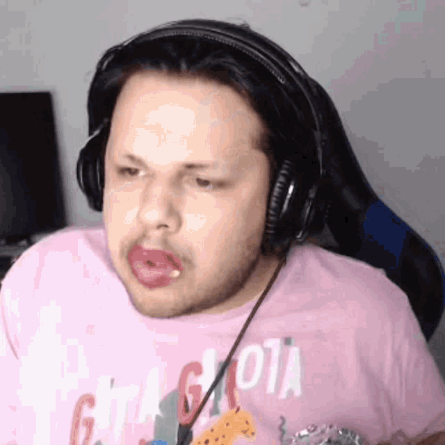 a man wearing headphones and a pink shirt is making a funny face while sitting in a chair .