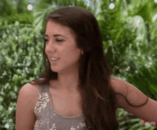 a woman wearing a sequined top is standing in front of a lush green forest .